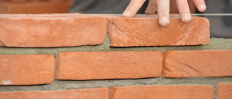 A bricklayer bedding a red facing brick onto a fresh mortar joint on a brick wall