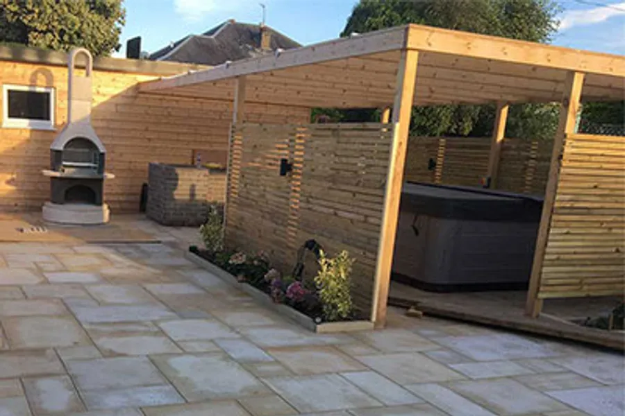 A hot tub under a timber shelter with slatted privacy screening on a sandstone patio base